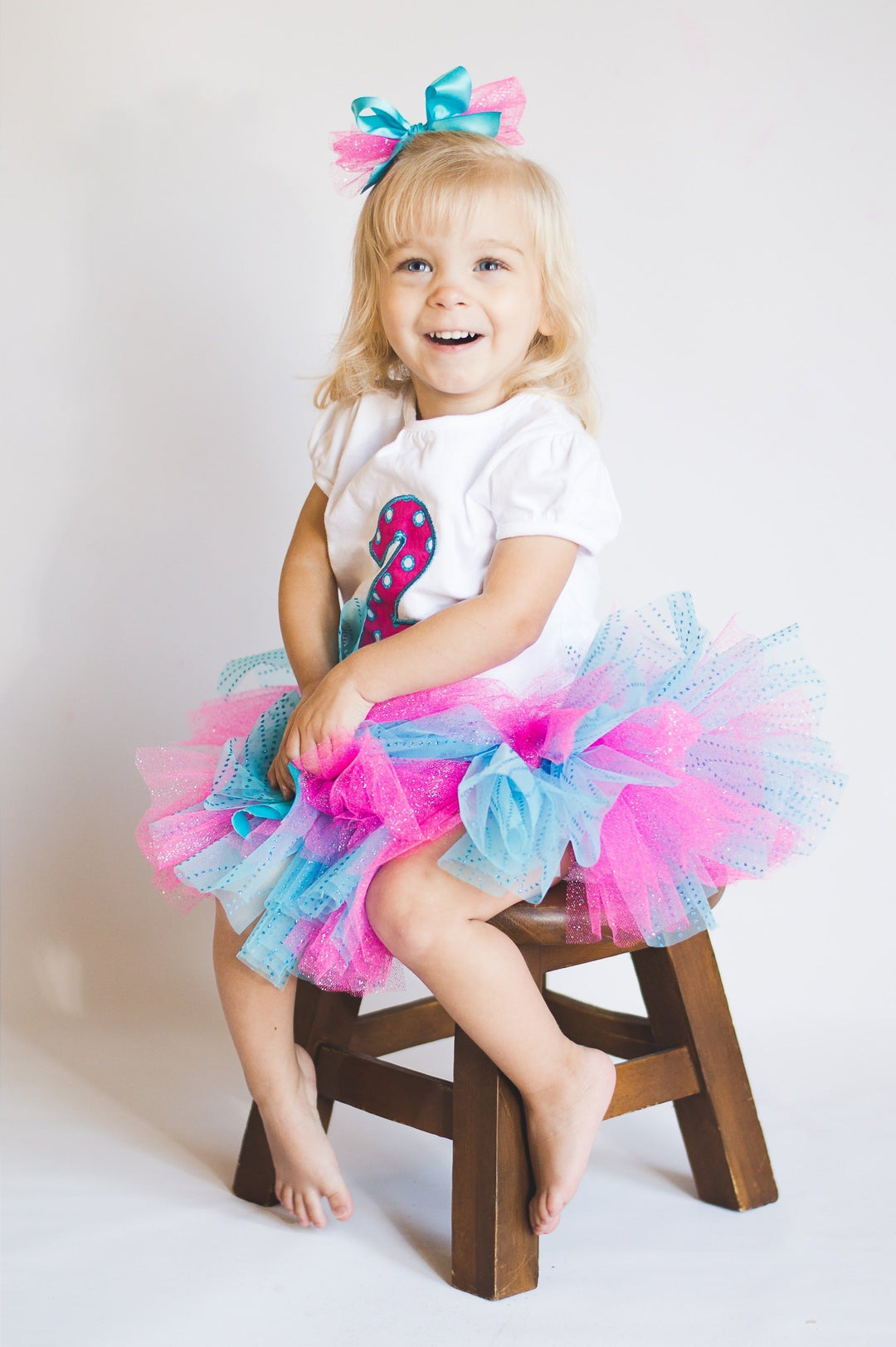 Smiling toddler wearing a pink and blue tutu and a birthday shirt with a number two appliqué, sitting on a wooden stool – personalized children’s birthday outfit by Pisgah Needleworks.