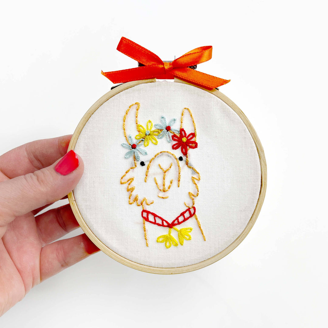 Embroidery hoop with a llama design and red bow held by a hand on a white background