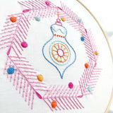 Embroidery hoop with colorful embroidered design featuring a decorative element and pink herringbone pattern.