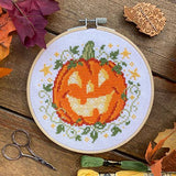 Cross-stitched pumpkin design in an embroidery hoop on a wooden surface with fall leaves and scissors.