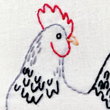 Embroidered chicken design on fabric with red comb and black tail feathers.