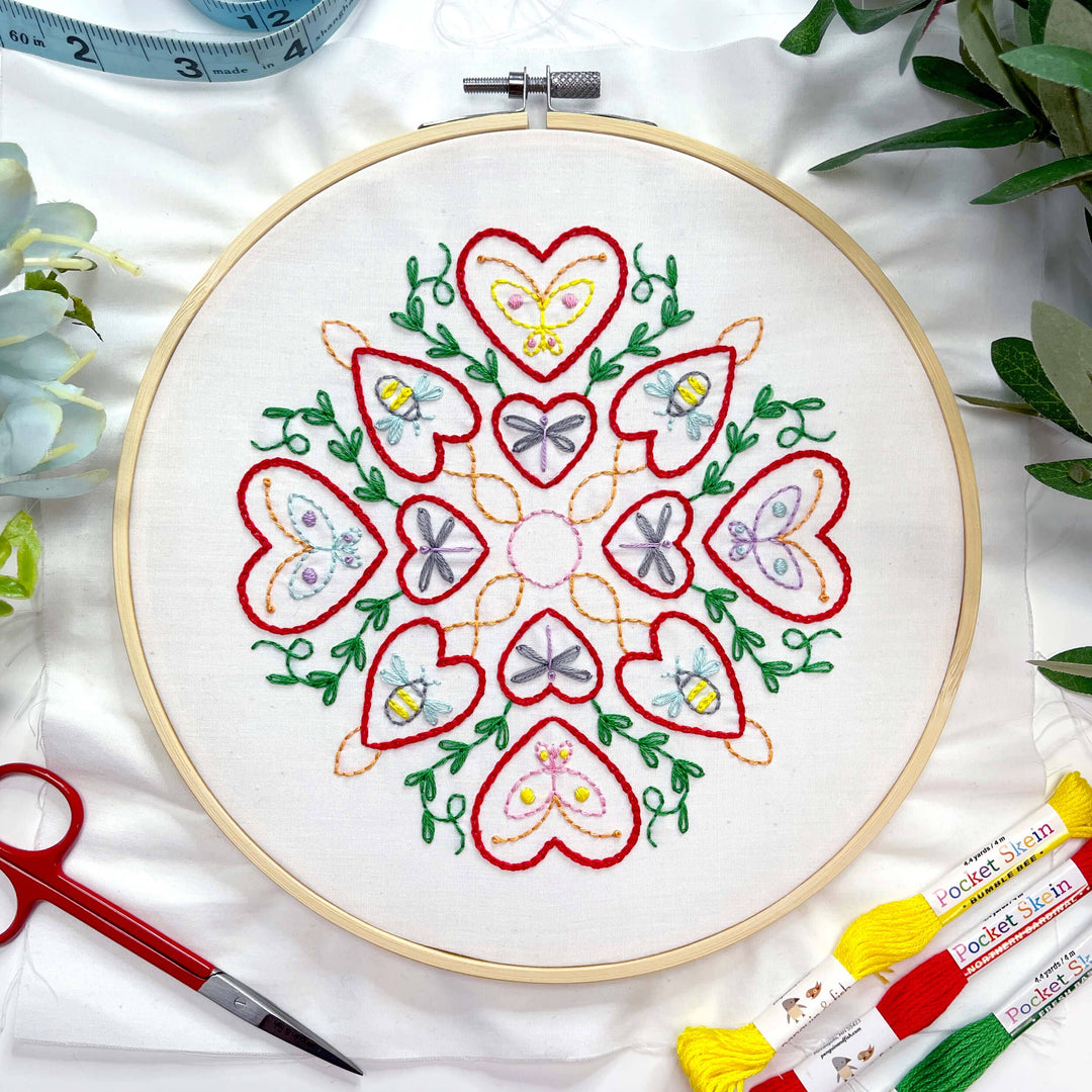Embroidery hoop with heart and butterfly design on white fabric, surrounded by embroidery supplies.