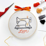 Embroidery hoop with a sewing machine design and 'love' text, surrounded by sewing materials on a white background.