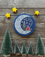 Embroidery hoop with a Christmas-themed design on a wooden surface with small trees and stars.