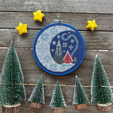 Embroidery hoop with a Christmas-themed design on a wooden background with small trees and stars.