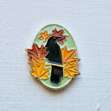 Pin with a black bird surrounded by colorful leaves on a textured white background
