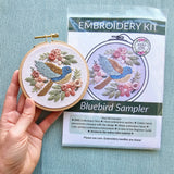 Embroidery hoop with bluebird design and embroidery kit packaging on a light blue background