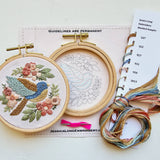 Embroidery hoops with floral and bird design, thread, and a pattern book on a white background.
