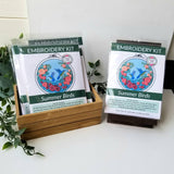 Embroidery kit packaging with 'Summer Birds' design on a white surface with greenery.