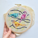 Embroidery hoop with colorful birds on a branch design held by a hand against a light background
