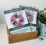 Two embroidery kits titled 'Floral Flourish' in a wooden holder with a plant in the background.