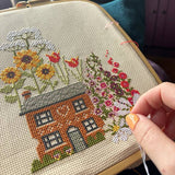 Cross-stitching a house with flowers on fabric, held by a person.