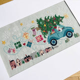 Cross-stitched panel with a blue truck carrying a Christmas tree and festive elements on a light background.