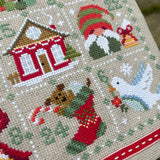 Cross-stitched Christmas scene with a house, gnome, dog, and stocking on a beige background.