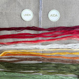 Collection of embroidery threads on a fabric background with labels for 'Linen' and 'Aida'.