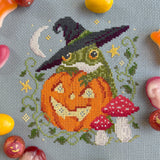 Cross-stitched design of a frog with a witch hat, pumpkin, and mushroom on fabric.