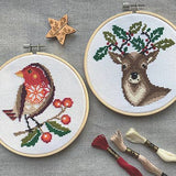Two cross-stitched pieces on hoops with a robin and deer design, surrounded by embroidery tools on a gray background.