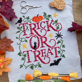 Cross-stitched Halloween design with 'Trick or Treat' text on fabric, surrounded by autumn leaves and embroidery supplies.