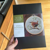 Cross stitch kit packaging with a wren and snowdrops design held by a hand on a wooden surface.