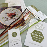 Cross-stitch kit with wren design, threads, and instructions on a green cutting mat.