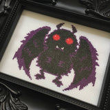 Cross-stitched creature with purple wings and red eyes on a white background, framed in black.