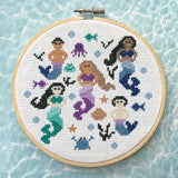 Cross-stitched design of mermaids and sea creatures in an embroidery hoop on a light blue water pattern background.