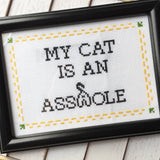 Framed cross-stitched text with a humorous message about a cat, on a wooden surface.