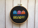 Cross-stitched design of Pac-Man ghosts and 'READY!' text in a wooden hoop on a light wood background.