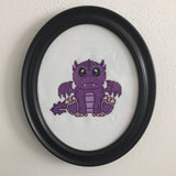 Framed cross-stitched purple dragon on a white background