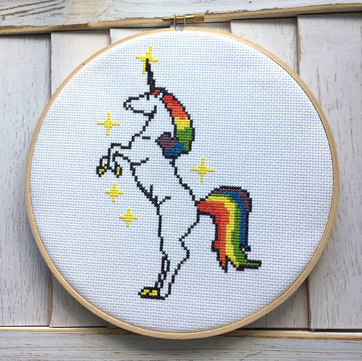 Cross-stitched unicorn with rainbow tail and horn in a wooden hoop on a light wooden background