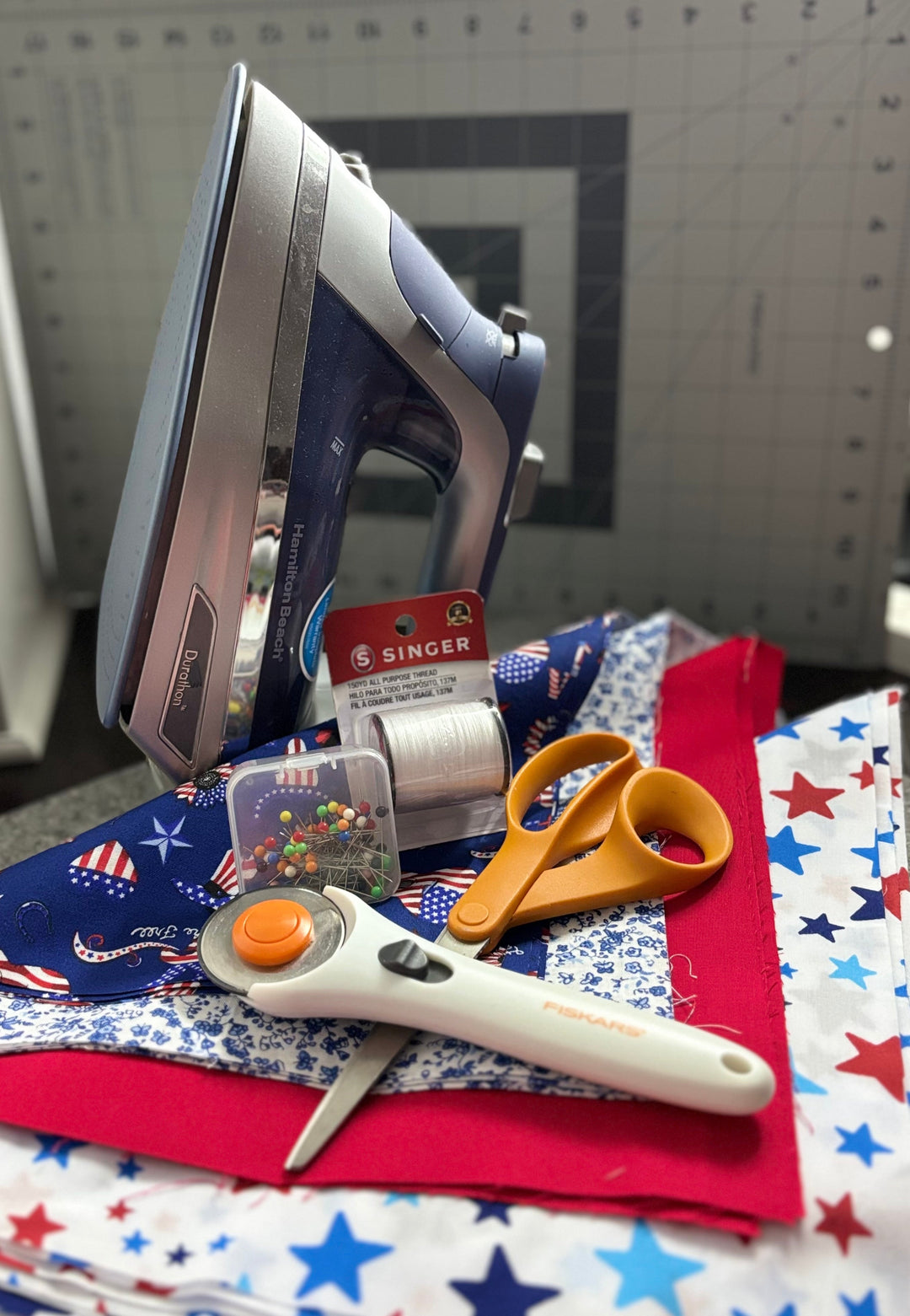 Beginner quilting supplies including a Hamilton Beach iron, rotary cutter, fabric scissors, sewing pins, thread, and patriotic-themed fat quarters on a cutting mat – Pisgah Needleworks quilting tools and fabrics.