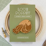 Wooden bird-shaped magnetic needle minder with packaging labeled 'Love Poppet' on a textured surface.