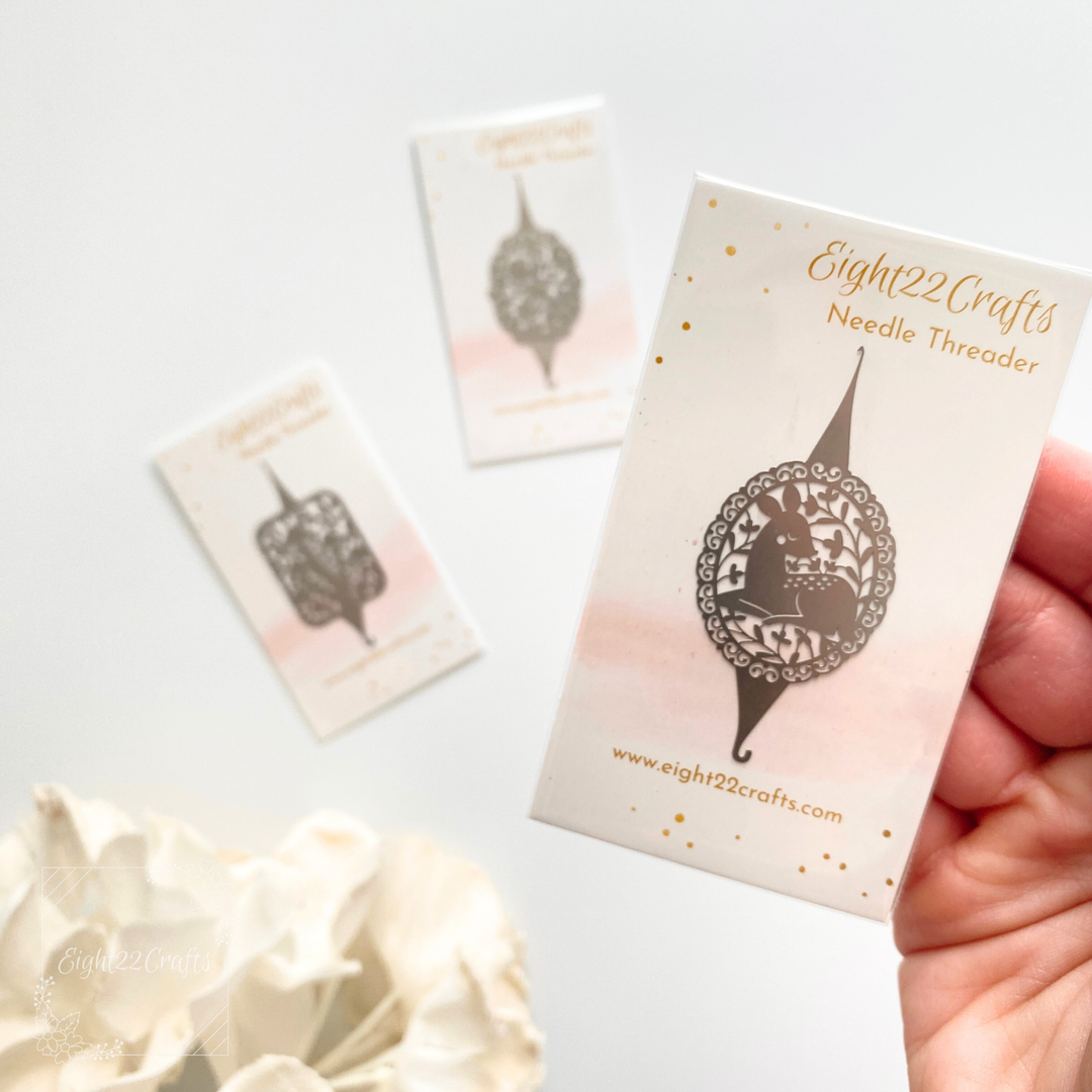 Hand holding a 'Eight22Crafts Needle Threader' packaging with floral background