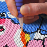 Hand using a bead placer to create a colorful pixel art design on a beaded canvas.