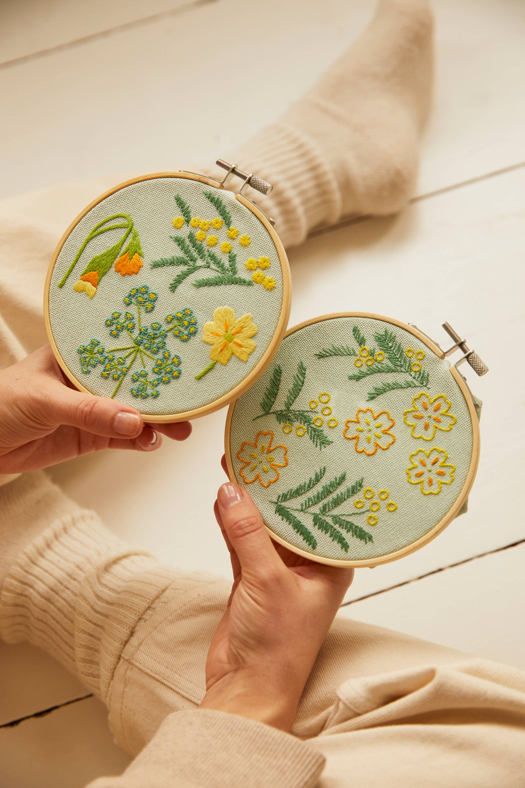 Two embroidery hoops with floral patterns held by a person wearing a beige sweater.