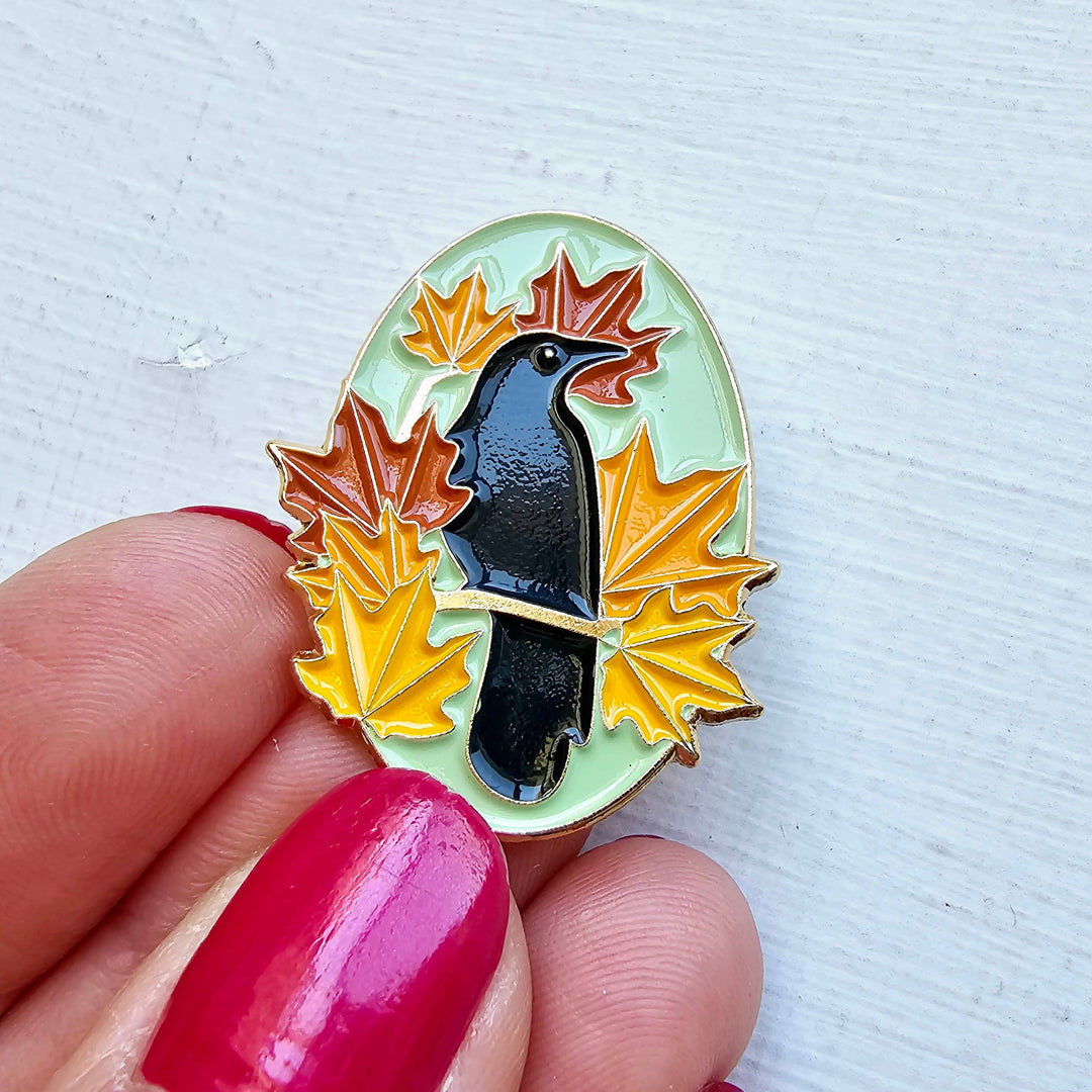 Pin with a bird and autumn leaves held by a hand with red nail polish on a light gray background