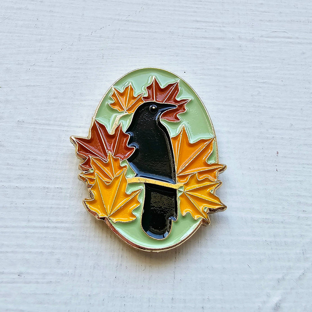 Pin with a black bird surrounded by colorful leaves on a textured white background