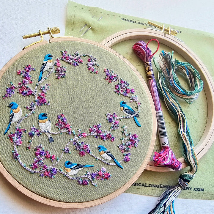 Embroidery hoop with bird and floral pattern on green fabric, with embroidery threads and tools.