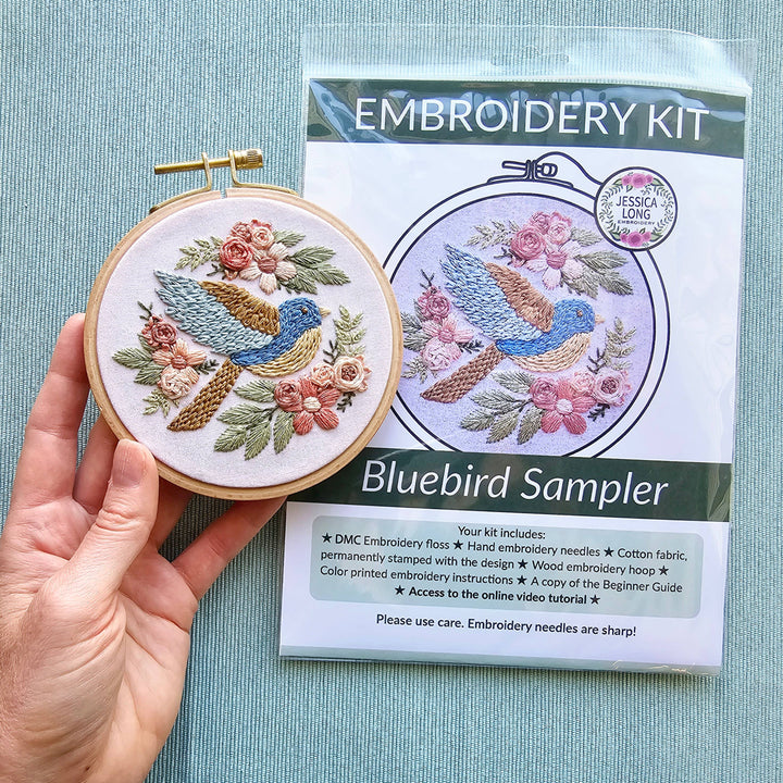 Embroidery hoop with bluebird design and embroidery kit packaging on a light blue background