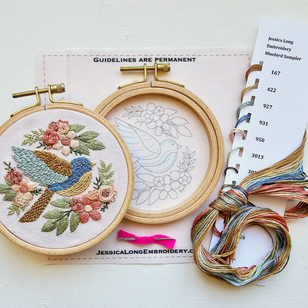 Embroidery hoops with floral and bird design, thread, and a pattern book on a white background.