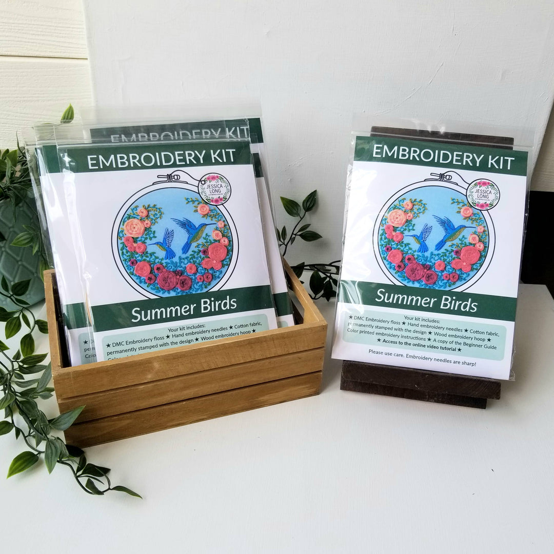 Embroidery kit packaging with 'Summer Birds' design on a white surface with greenery.