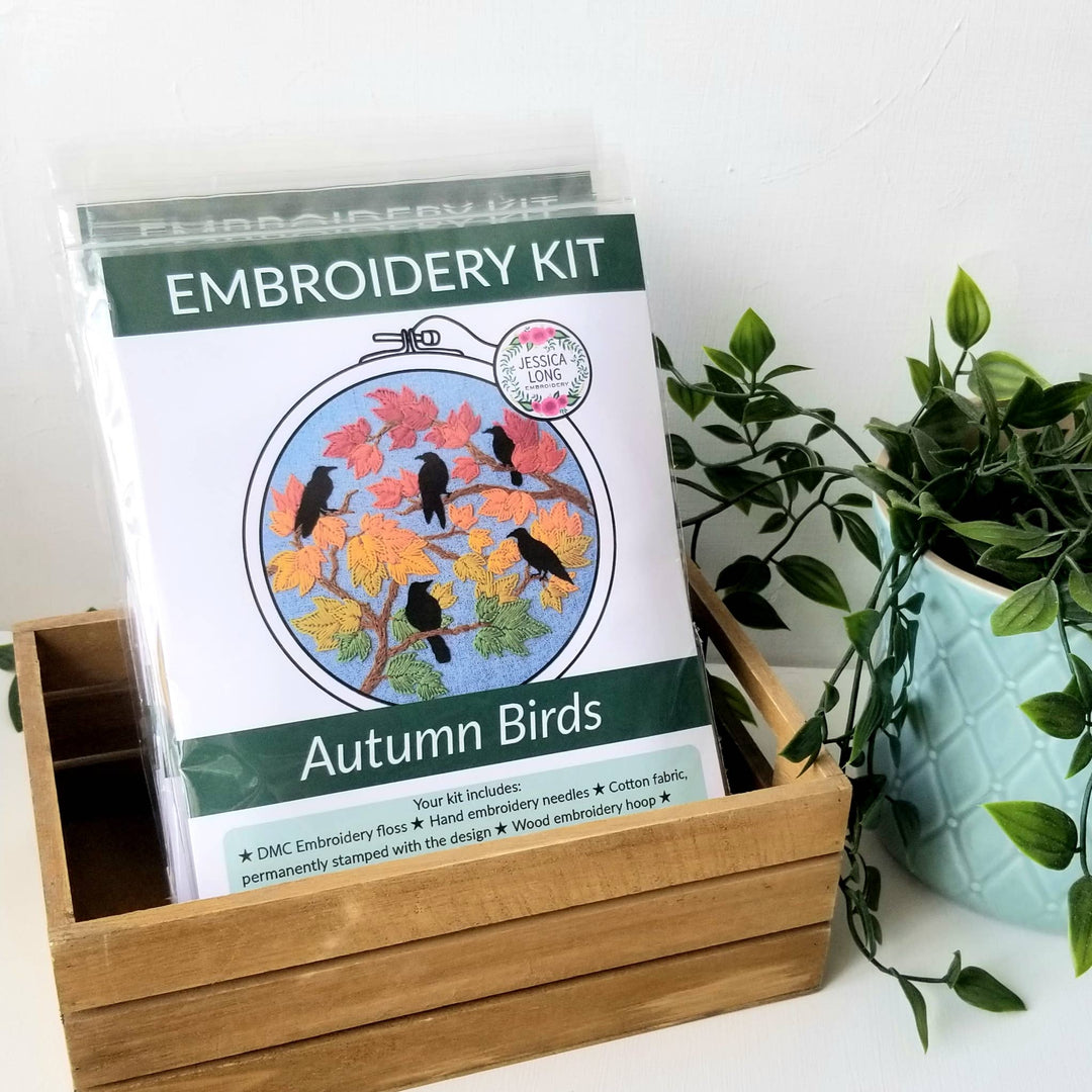 Embroidery kit for 'Autumn Birds' design in a wooden box with a plant in the background.