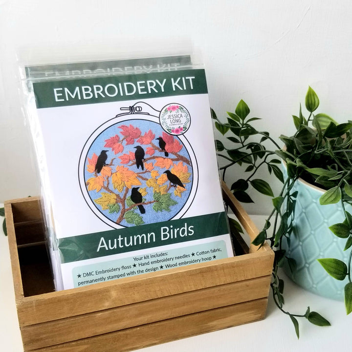 Embroidery kit for 'Autumn Birds' design in a wooden box with a plant in the background.