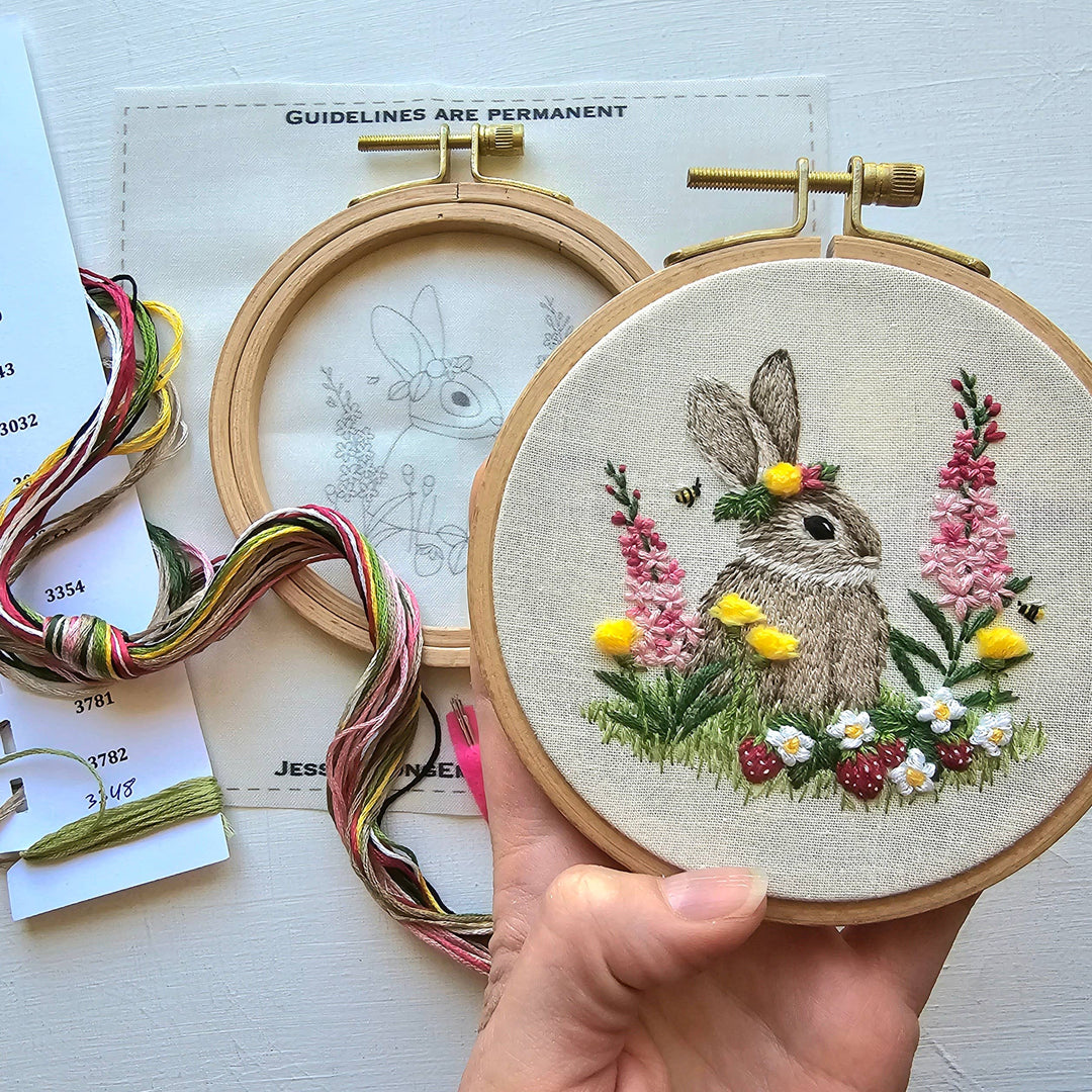 Embroidery hoop with a rabbit and floral design held by a hand, with embroidery threads and patterns in the background.
