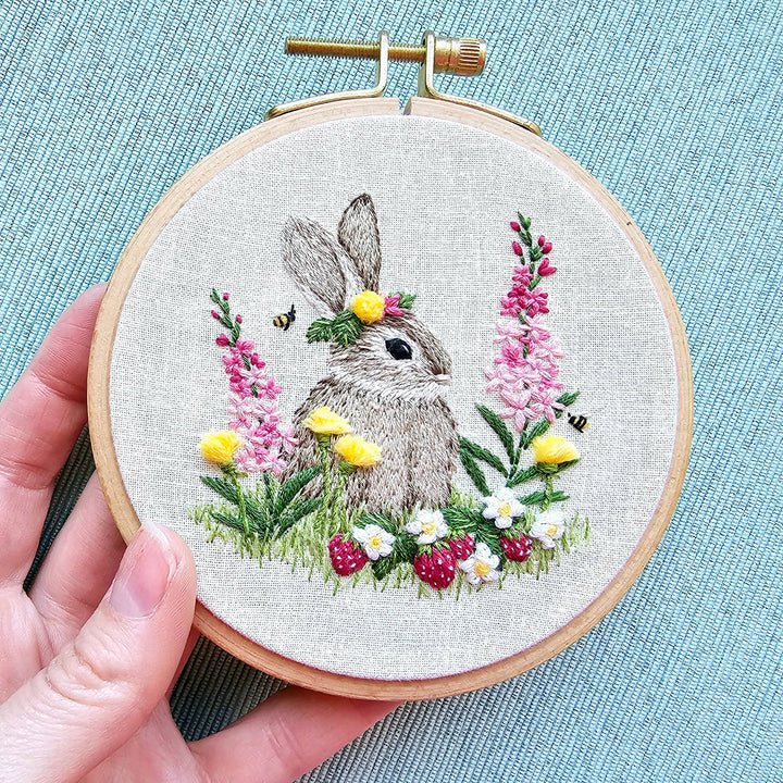 Embroidery hoop with a rabbit and floral design held by a hand on a textured blue background.
