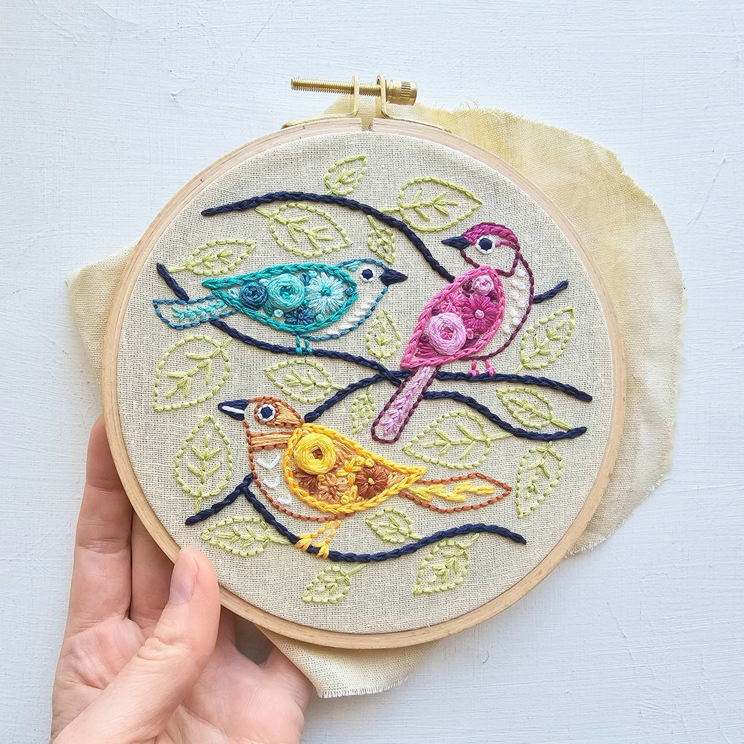 Embroidery hoop with colorful birds on a branch design held by a hand against a light background