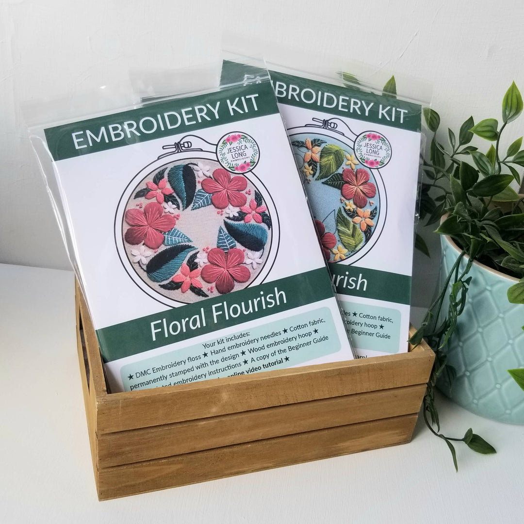 Two embroidery kits titled 'Floral Flourish' in a wooden holder with a plant in the background.