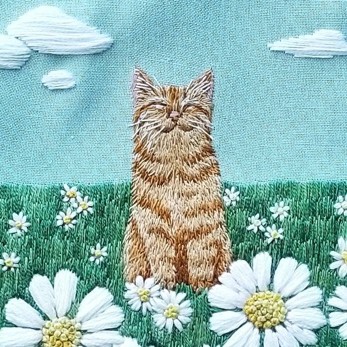 Embroidered design of a cat sitting in a field of flowers with a blue sky.