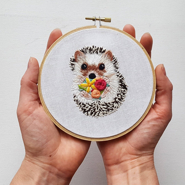 Embroidery of a hedgehog holding flowers in a wooden hoop held by hands against a light background