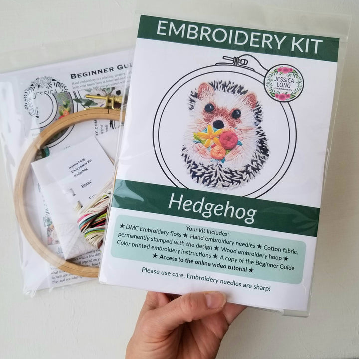 Embroidery kit for hedgehog design held by a hand, with an embroidery hoop and materials in the background.